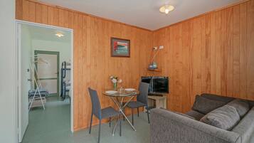 Un espace de vie compact avec un mur en bois, un canapé gris, une petite table à manger avec des chaises, un téléviseur et un tableau accroché au mur.