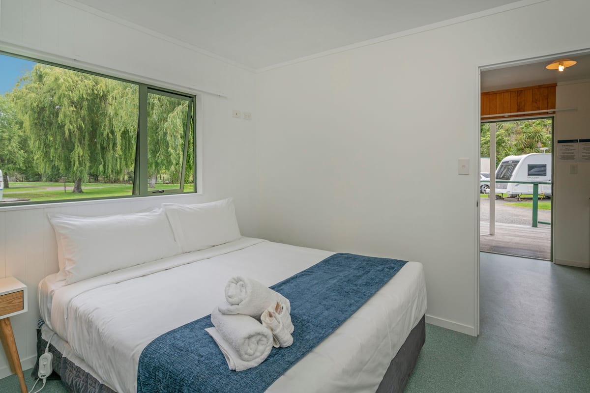 Habitación de hotel con cama, ventana con vista a árboles y puerta que da a un espacio exterior.
