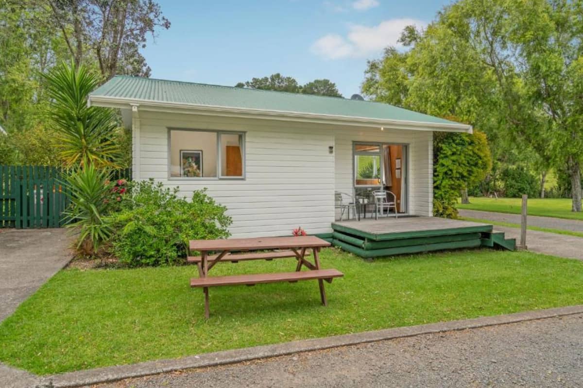 Una casa pequeña de un piso, con techo verde, un porche y una mesa de picnic.
