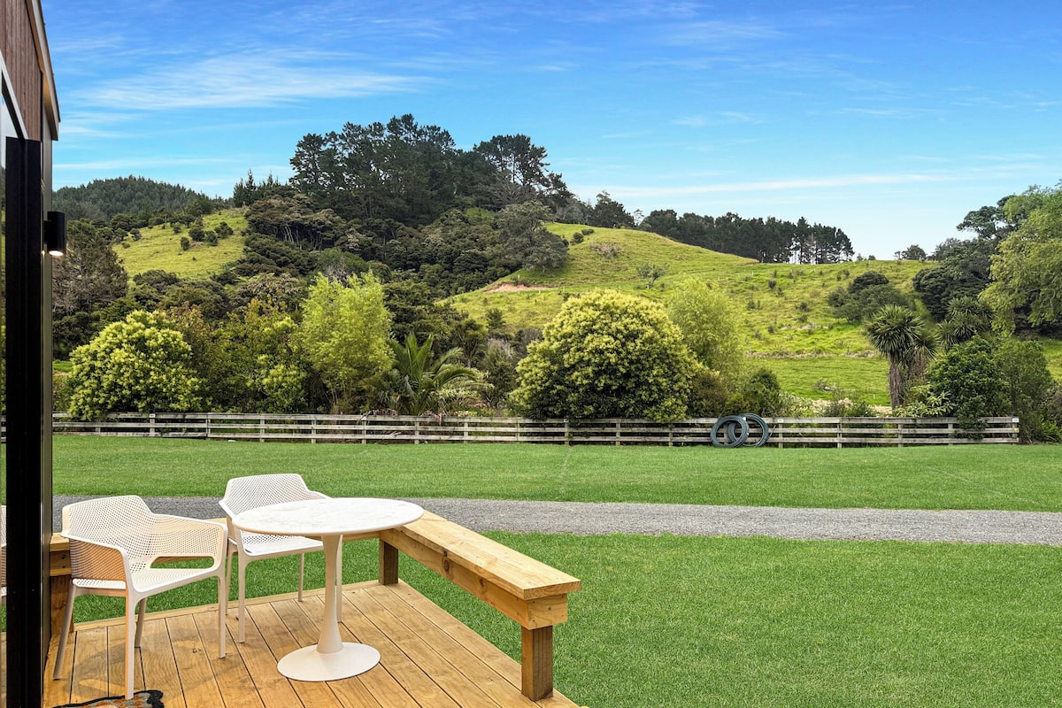 Una terraza de madera con una mesa, sillas, un banco y vistas a una colina cubierta de pasto y árboles.
