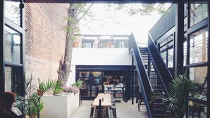 An outdoor cafe area with a long wooden table and chairs, surrounded by potted plants and a staircase leading to an upper level.