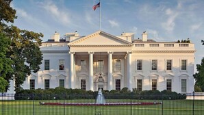 A large, white building with a central portico and columns, a flag on top, and a well-maintained lawn with a fence in front.