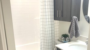 A bathroom with a white tiled wall, a white toilet, a bathtub with a white shower curtain, dark wooden cabinets, and a white sink. - ★Home away from Home ★ (Tremonton)