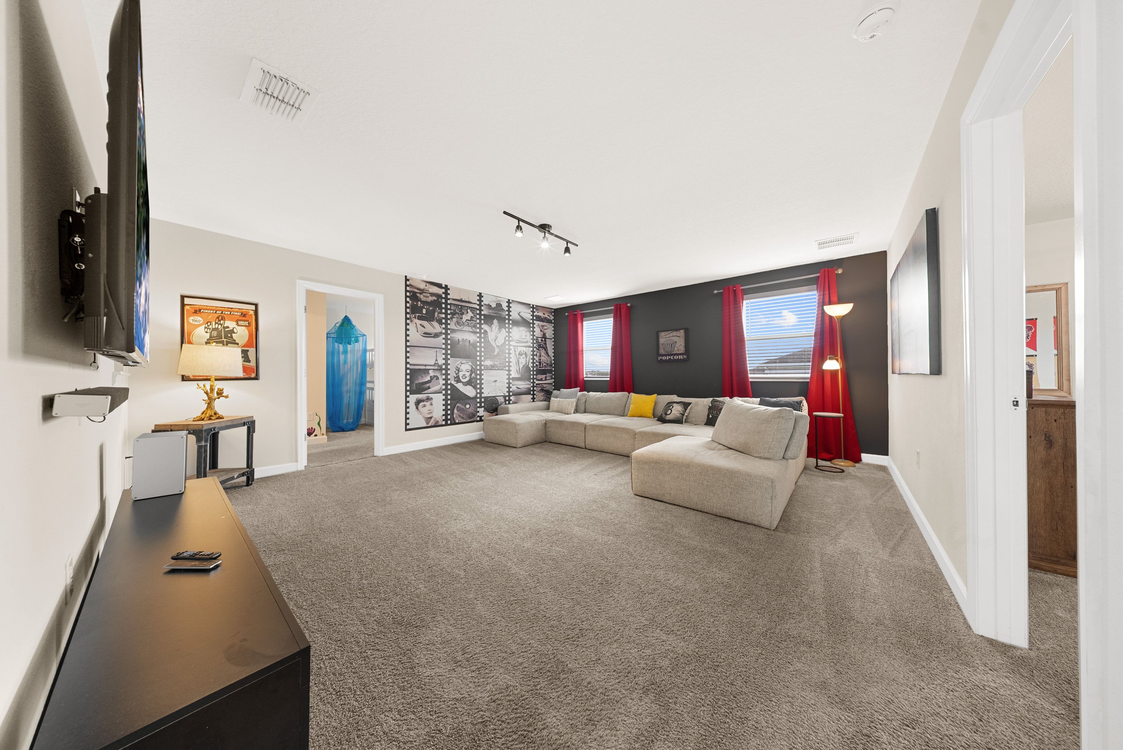 Un salon moderne avec un canapé d’angle, un téléviseur à écran plat fixé au mur et une grande fenêtre ornée de rideaux rouges.