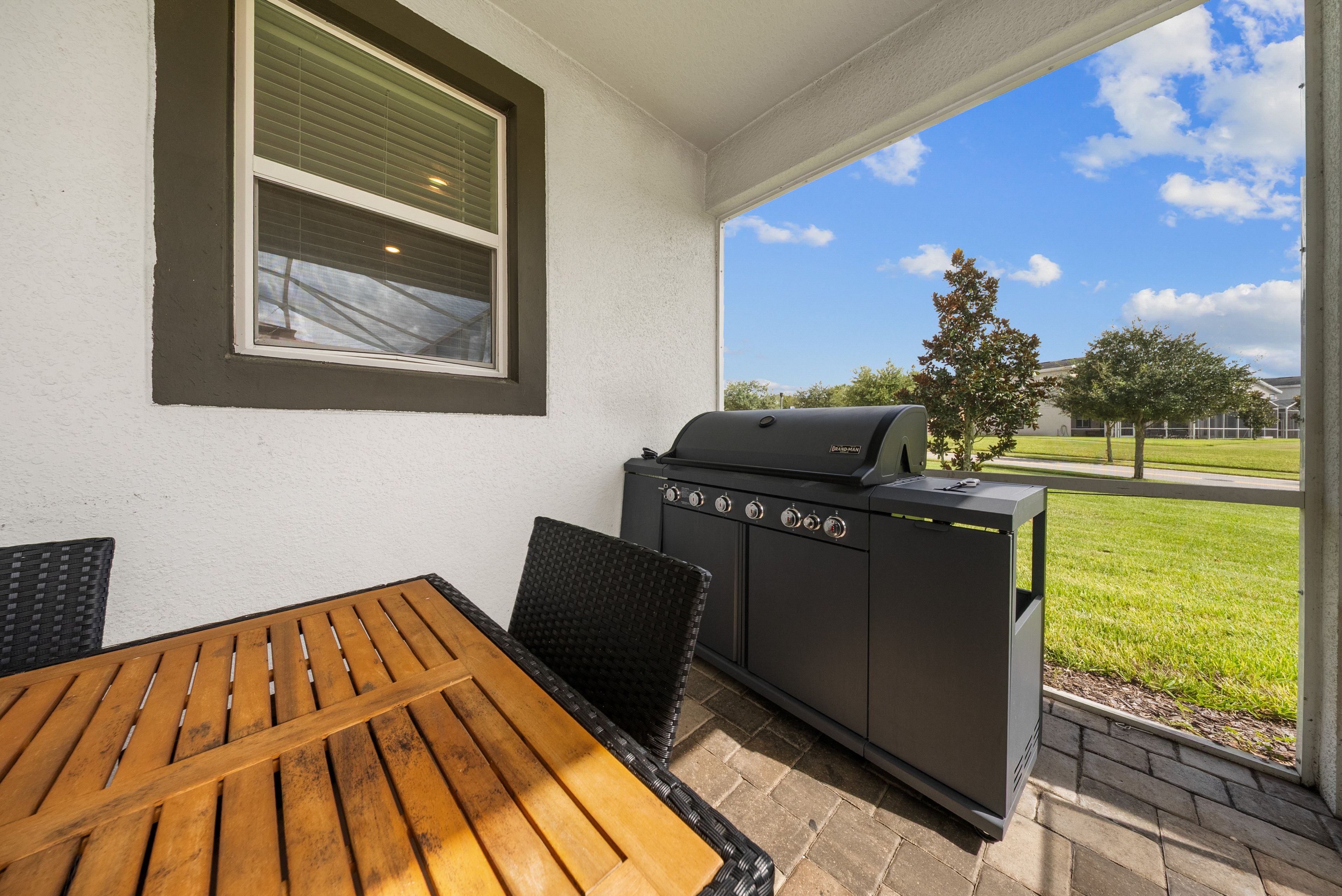 Un patio exterior con mesa y sillas de madera, una parrilla y vistas a un área verde y árboles.