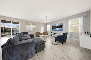 A spacious living room with a grey sectional sofa, a coffee table, a TV mounted on the wall, and a large window with blinds.