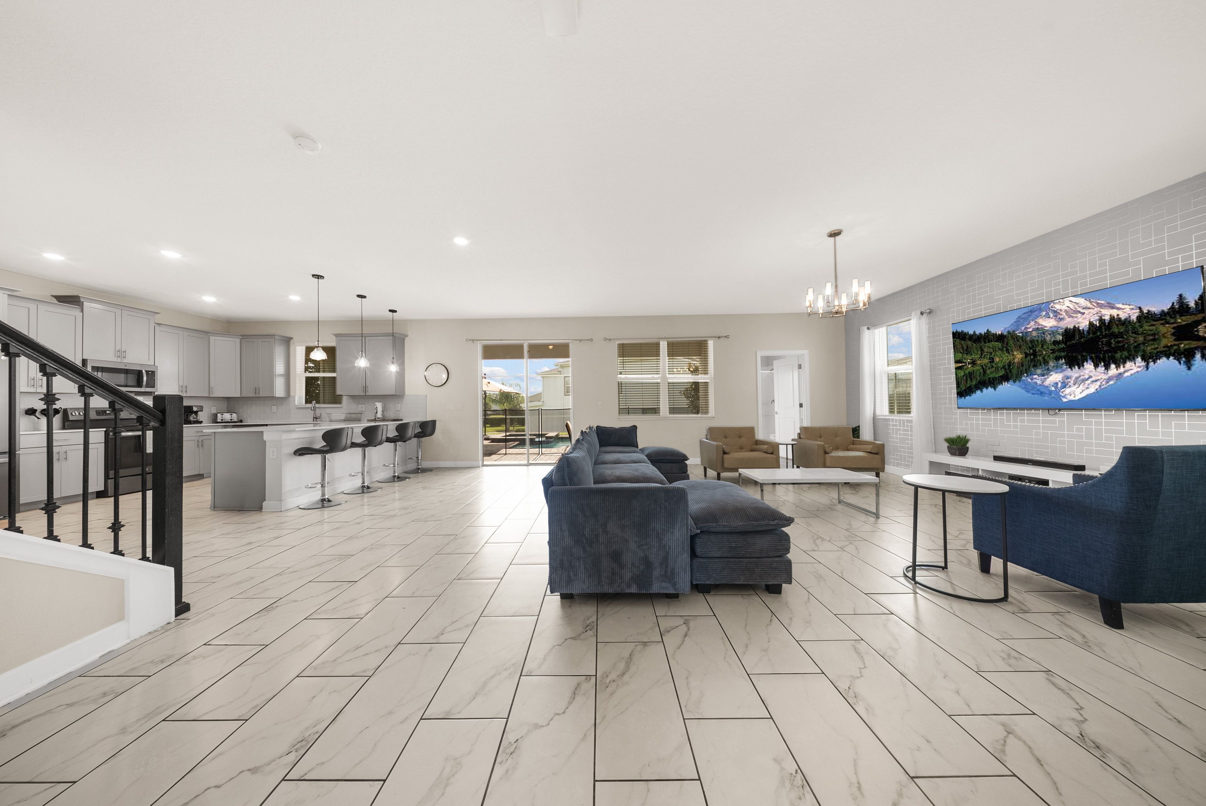 A spacious living area with a large sofa, a dining area with bar stools, a kitchen with modern appliances, and a large TV mounted on the wall.
