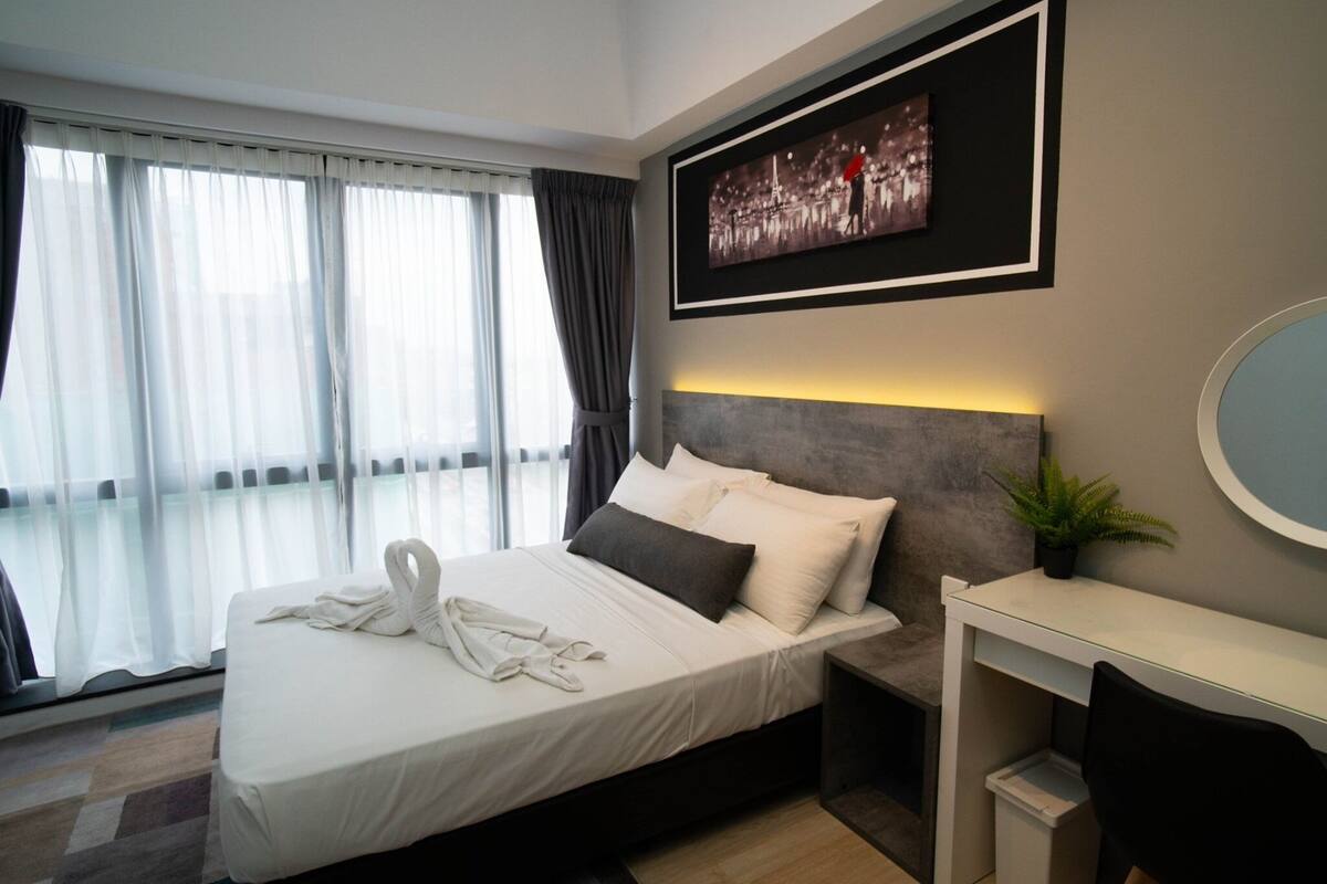 A modern hotel room with a large bed, a bedside table with a mirror, a framed picture on the wall, and a window with sheer curtains.