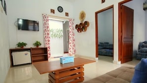 A living room with a wooden table, a sofa, a TV, a small fridge, and a door leading to another room.