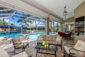 A spacious outdoor area with a pool, comfortable seating, and large windows offering views of palm trees and a clear sky. - Bailey Rose - Luxury Home Sailboat Access Canal (Cape Coral)