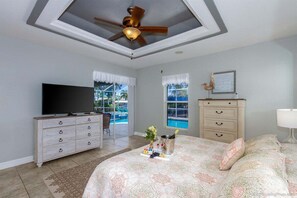A bedroom with a bed, a TV, a dresser, a ceiling fan, and a window with a view of a pool. - Bailey Rose - Luxury Home Sailboat Access Canal (Cape Coral)