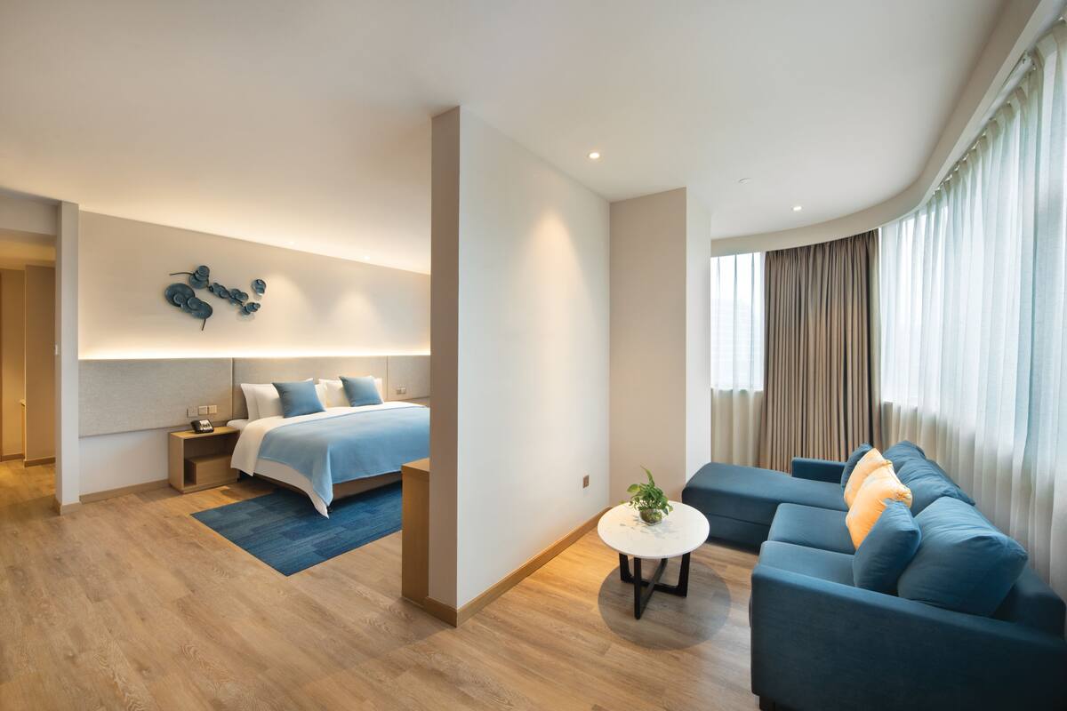 A modern hotel room with a bed, a sofa, a small table, and a wall-mounted artwork.