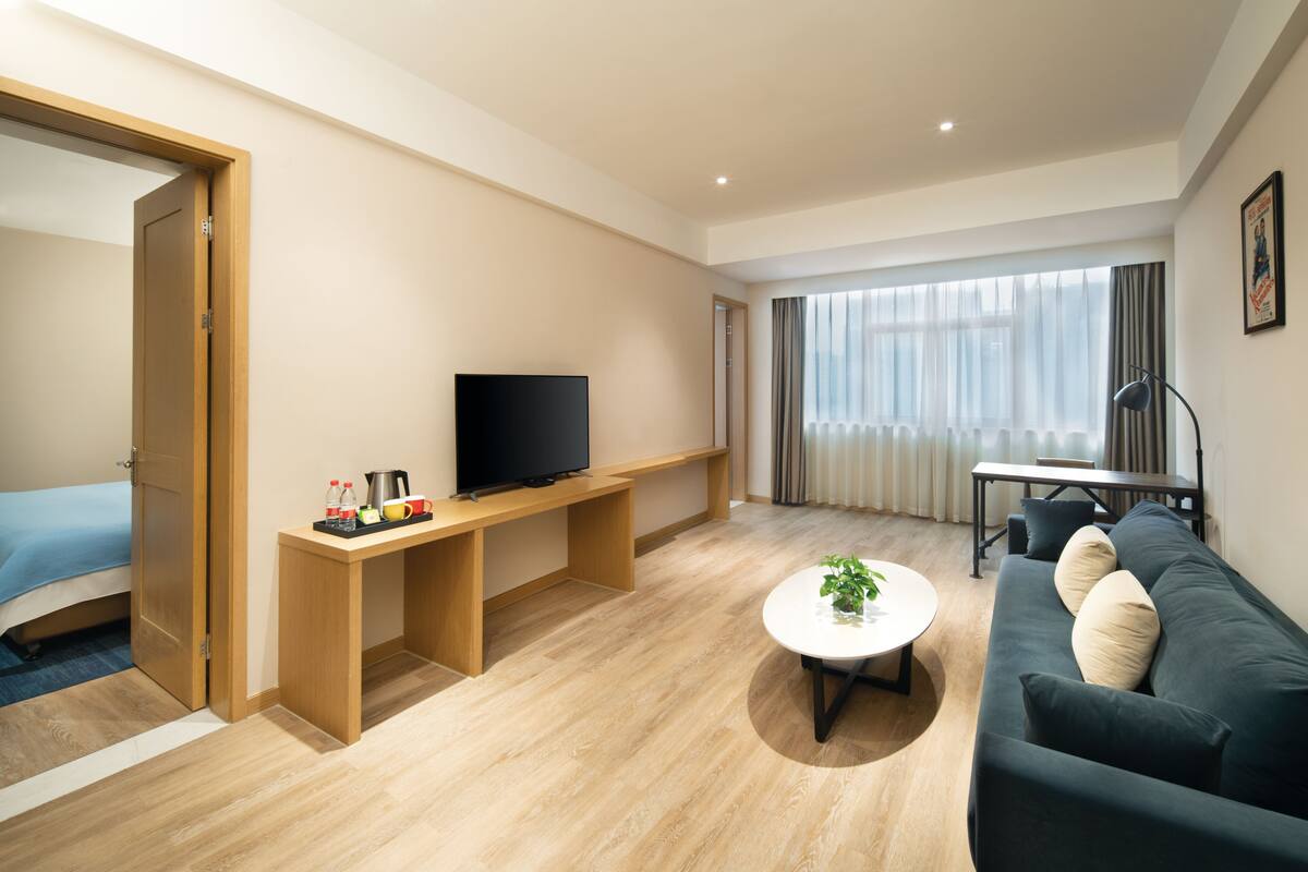 A modern hotel room with a wooden floor, a sofa, a TV on a wooden stand, a small table with a plant, and a bed with a blue bedspread.