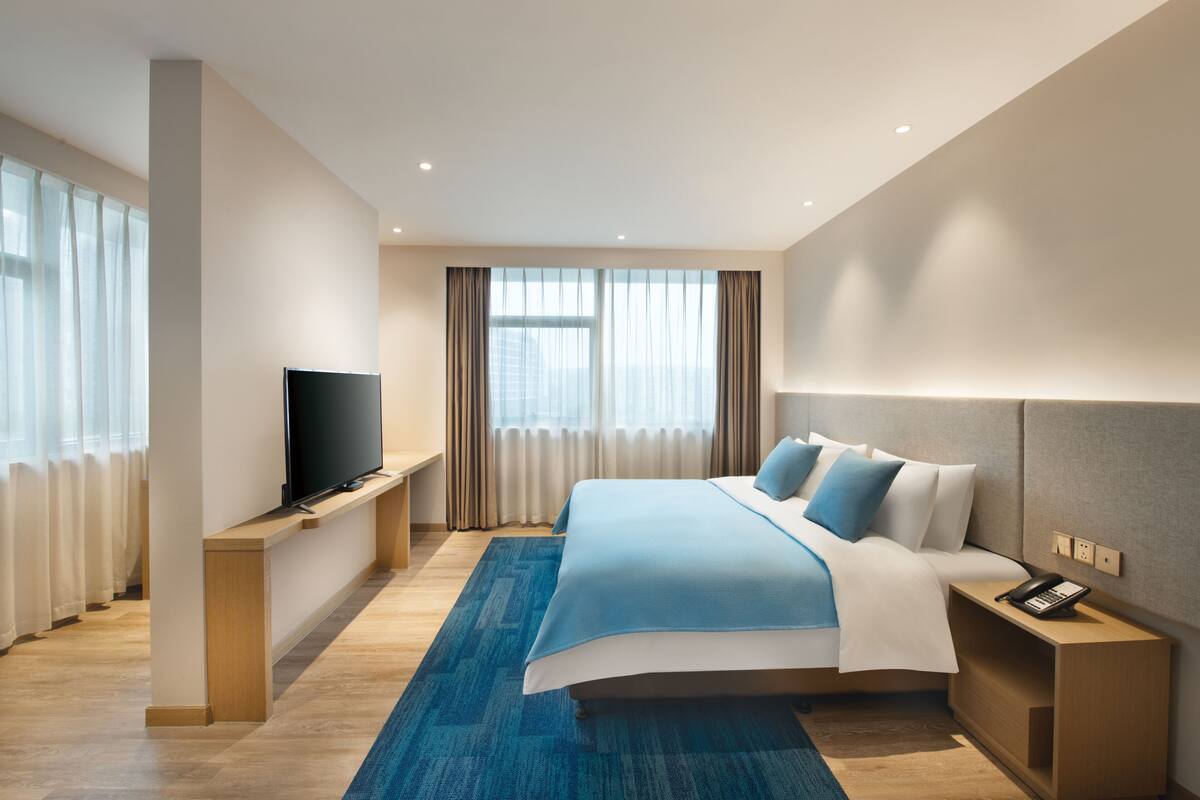A modern hotel room with a large bed, a flat-screen TV on a wooden console, a bedside telephone, and a view of the city through large windows.