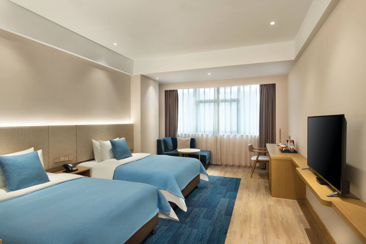 A hotel room with two beds, a desk, a TV, and a window with curtains.