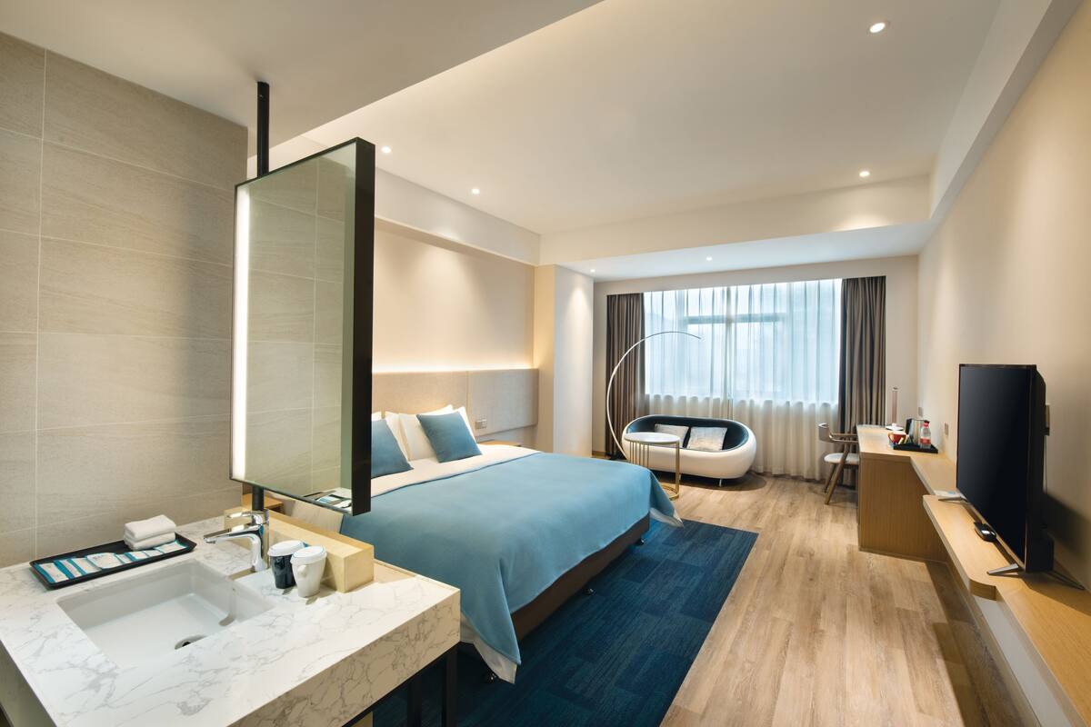A modern hotel room with a large bed, a separate seating area, a TV, and a bathroom with a marble sink.