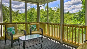 Un espace extérieur abrité, aménagé avec des meubles en osier et une table en verre, entouré d’une balustrade en bois offrant une vue sur les arbres verdoyants.