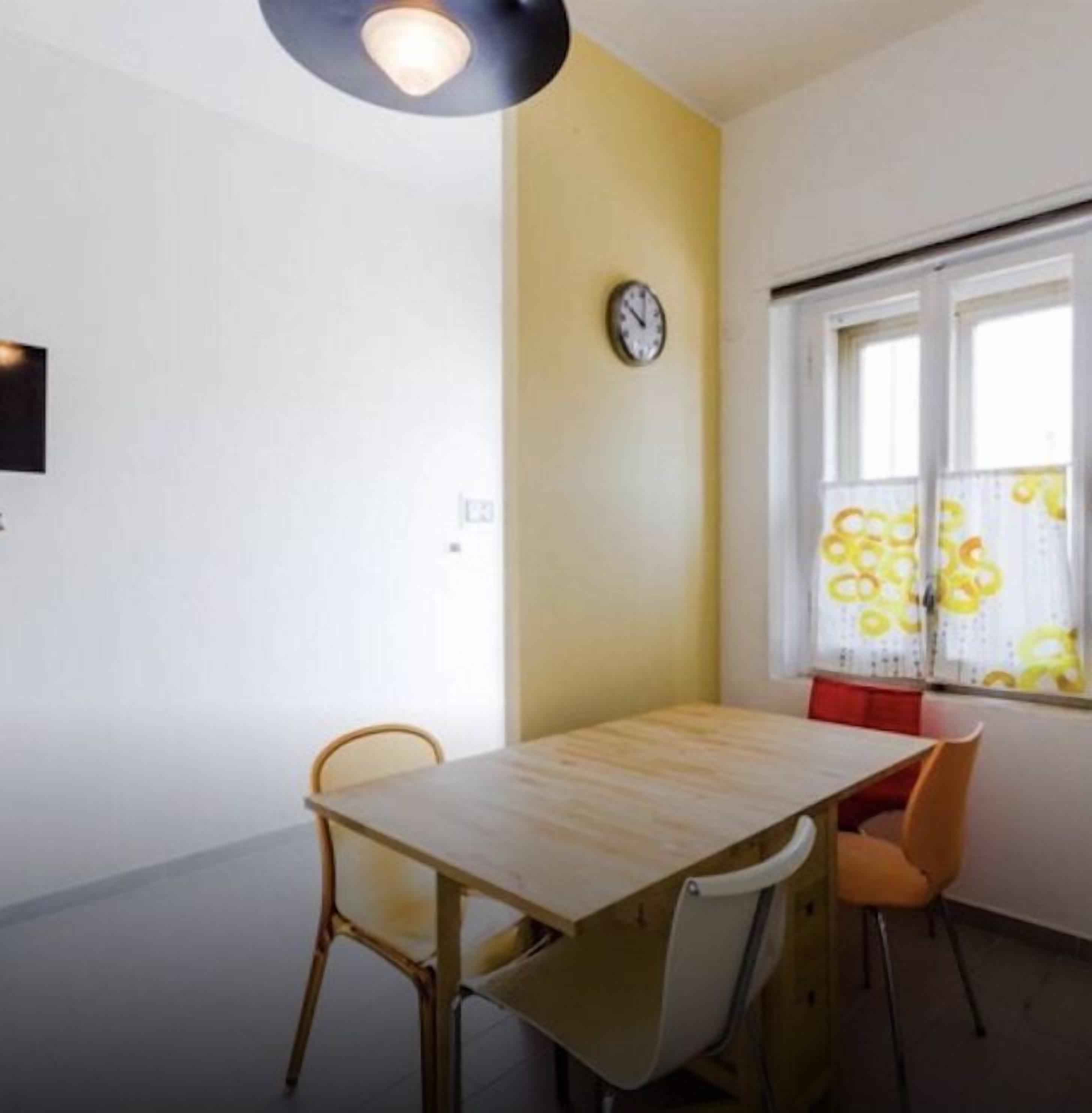 A dining area with a wooden table and chairs, a clock on the wall, and a window with a view of buildings.