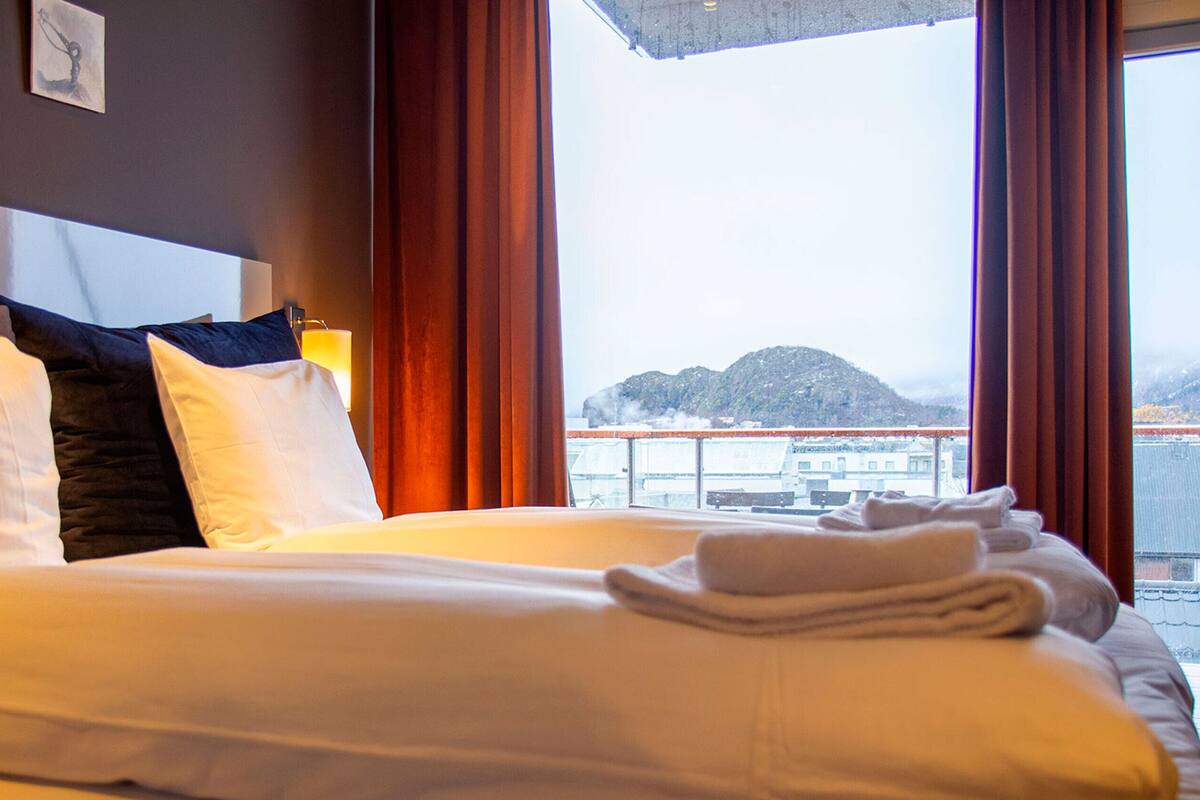 A hotel room with a bed, pillows, a nightstand, and a view of a body of water and mountains.
