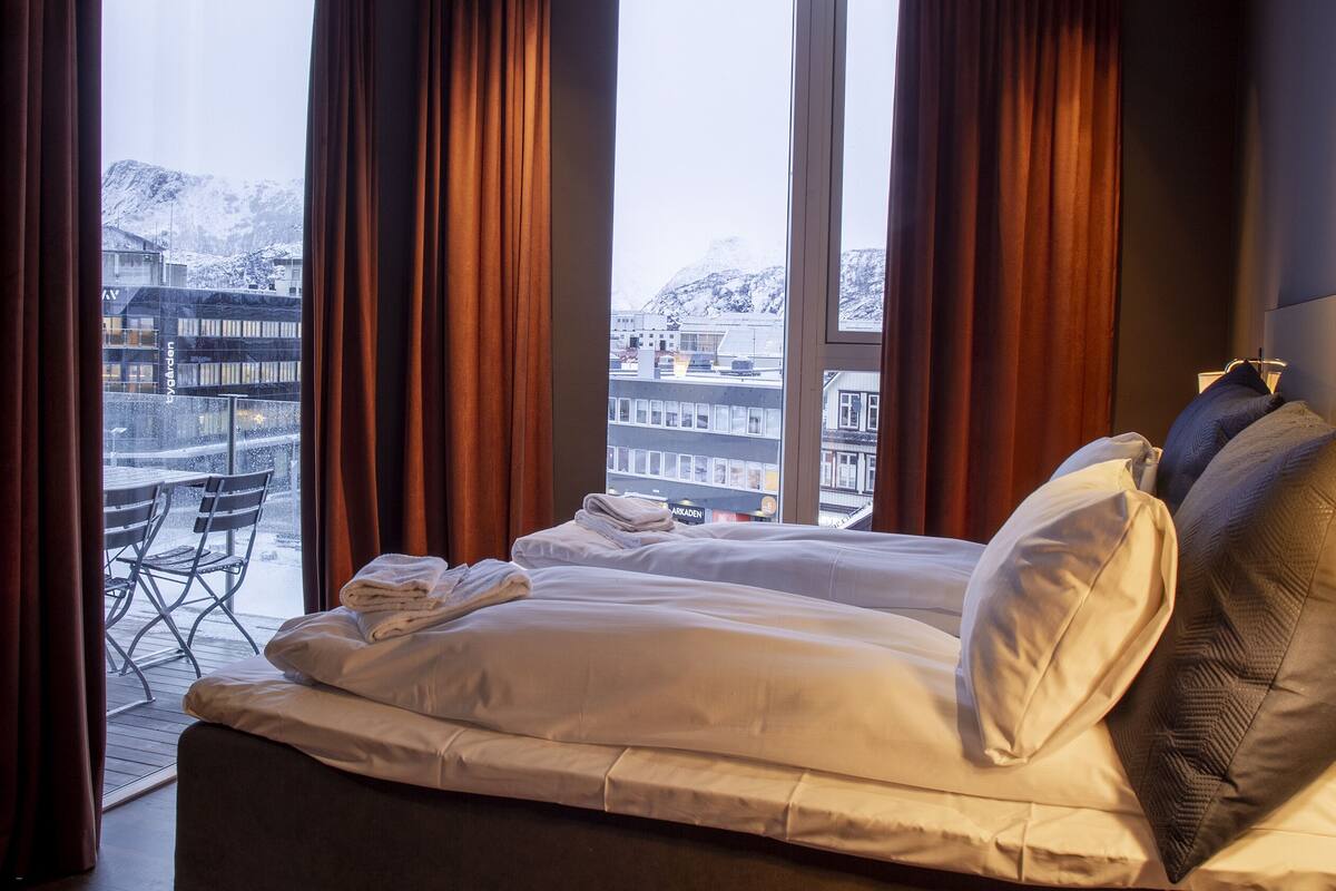 A hotel room with a bed, a chair, a table, and a view of a snowy mountain and buildings.