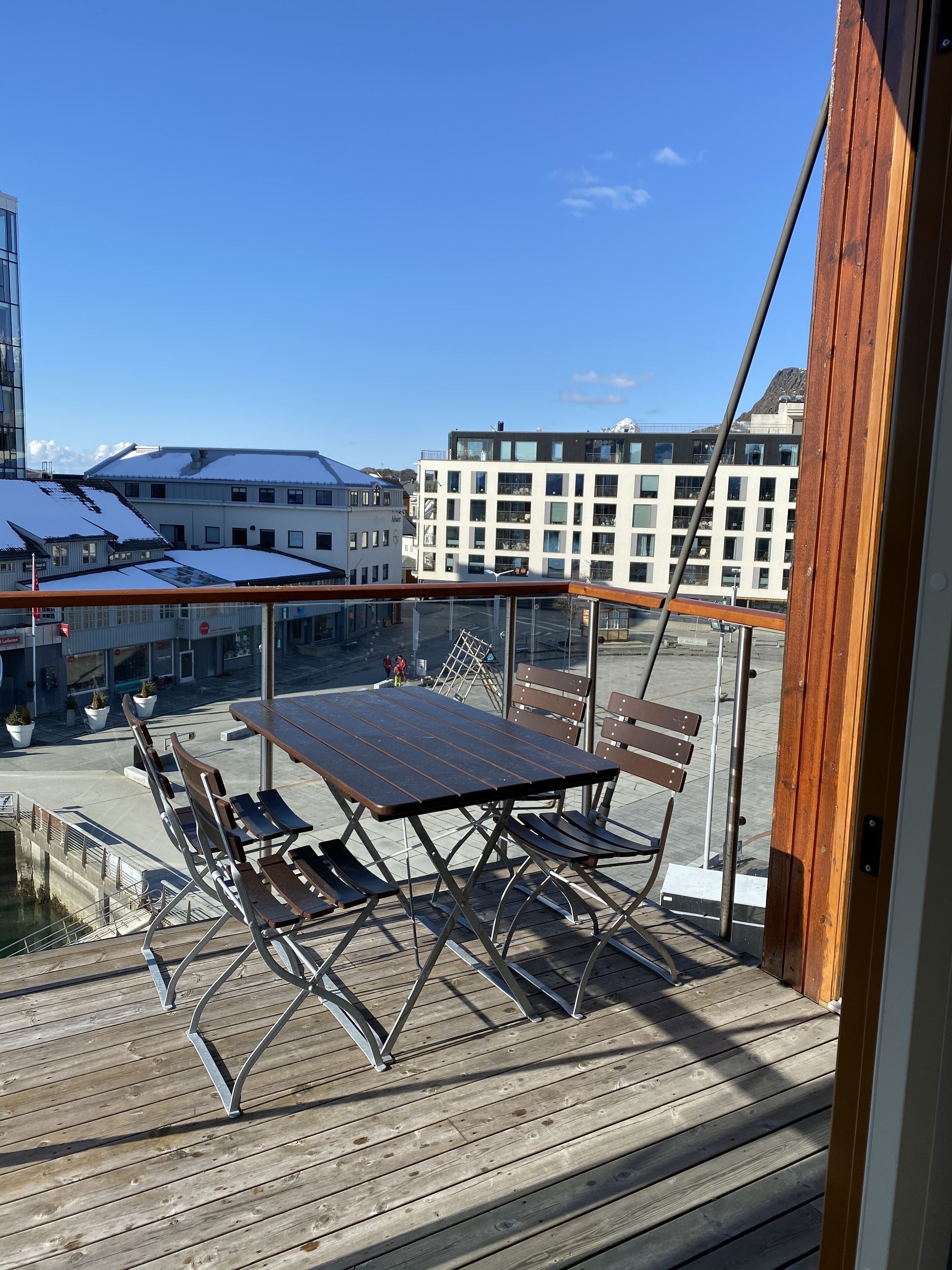 A balcony with a wooden table and chairs, overlooking a snowy urban area with buildings and a mountain in the background.