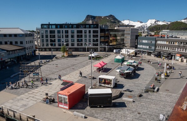 Una plaza pública con un edificio moderno, un paradero de autobús rojo y una montaña al fondo.