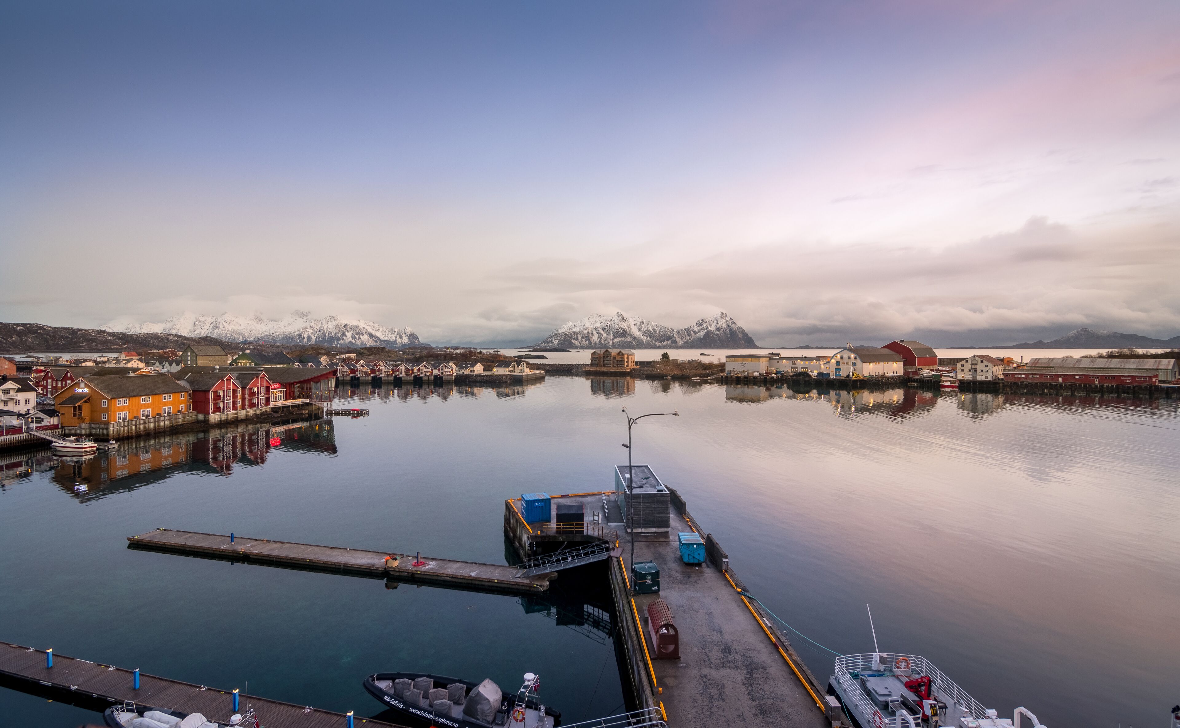 Un puerto con barcos amarrados, un muelle y edificios con montañas nevadas al fondo.
