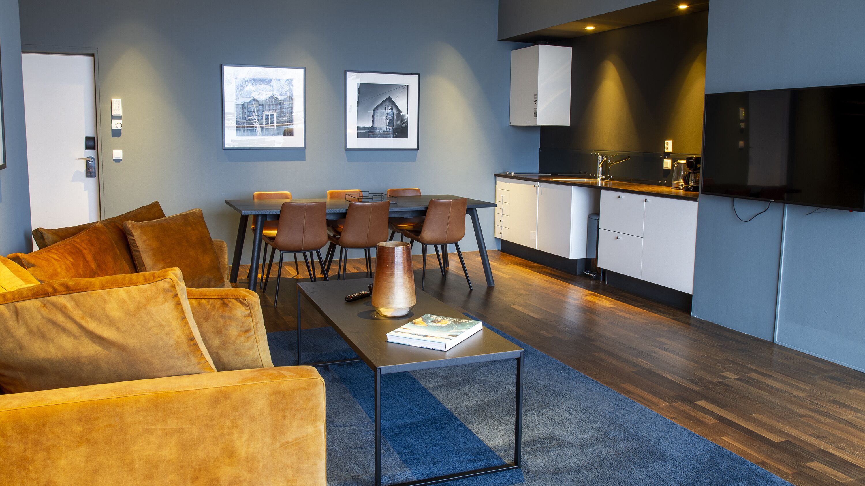 A modern living room with a sofa, coffee table, and a dining area with chairs. There is a kitchen area with cabinets and a TV mounted on the wall.