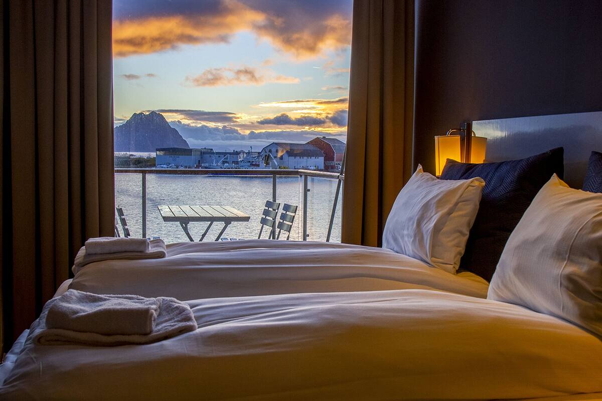 A hotel room with a bed, pillows, and a view of a dock and mountains.