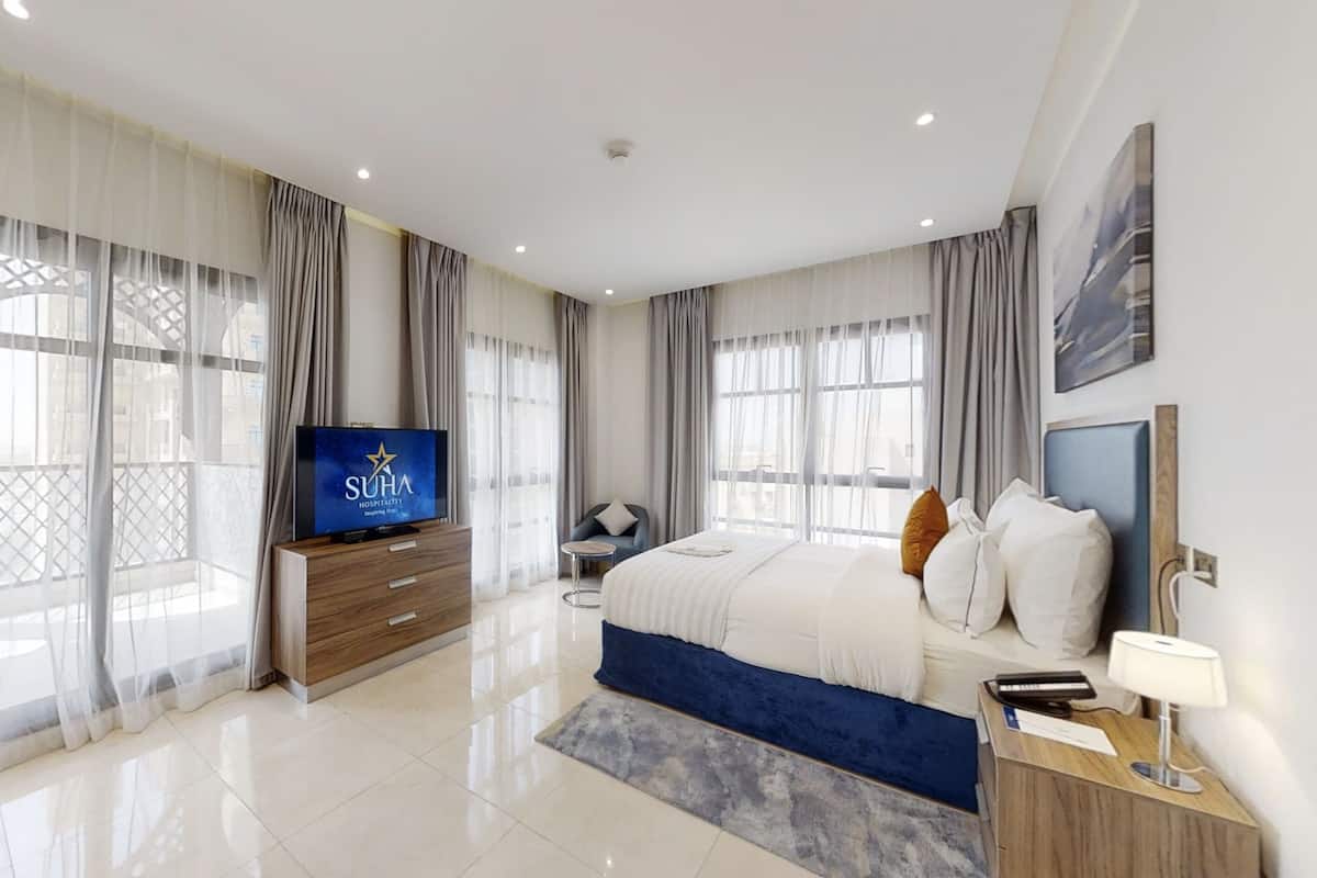 Una habitación de hotel moderna con una cama grande, un televisor sobre un aparador de madera y vistas de la ciudad a través de amplios ventanales.