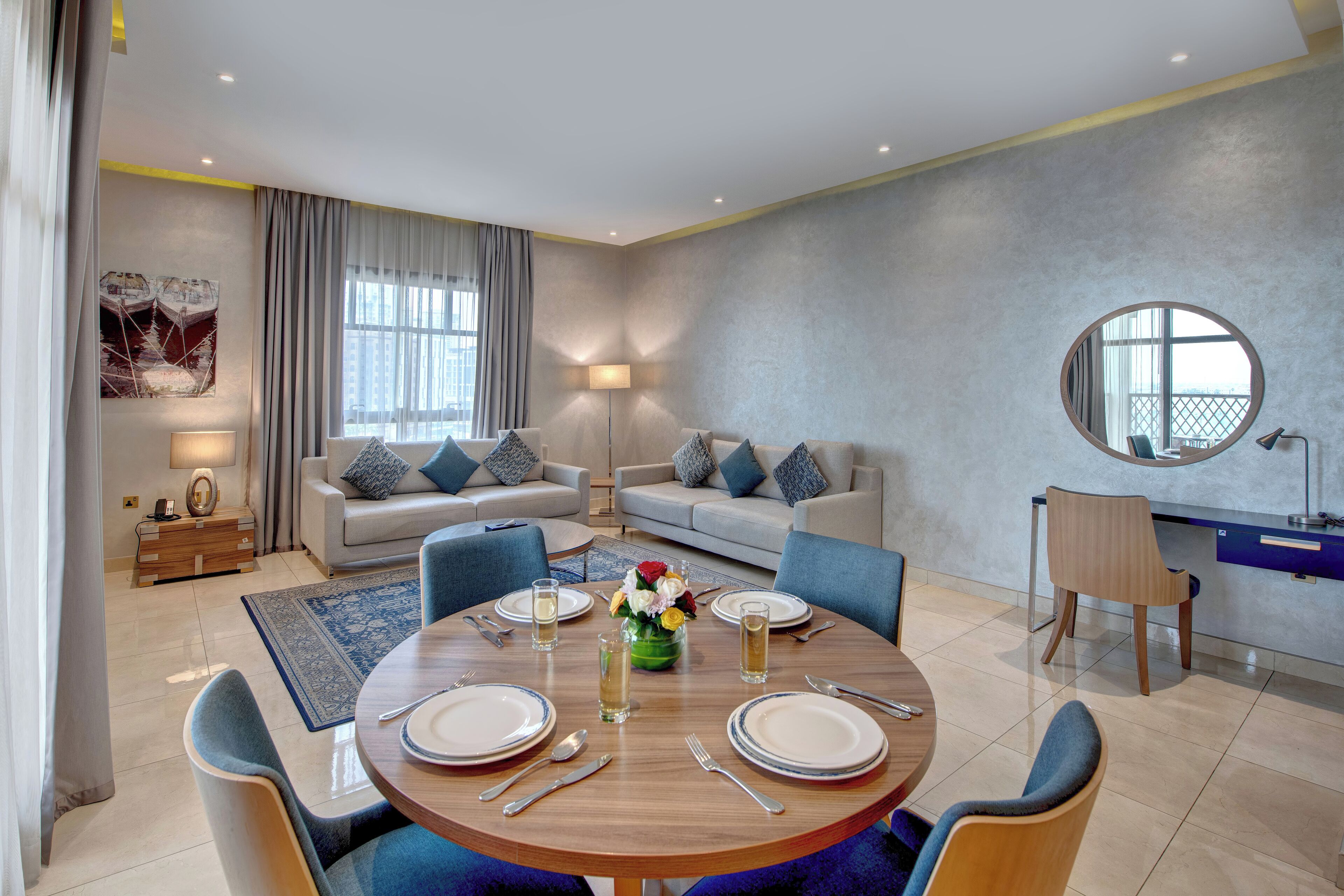 A modern living room with a dining area, featuring a round table set for four, two sofas, a coffee table with flowers, and a large window with curtains.