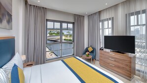 Une chambre d’hôtel moderne avec un grand lit, un téléviseur posé sur une commode en bois, une chaise et un balcon offrant une vue sur la ville.