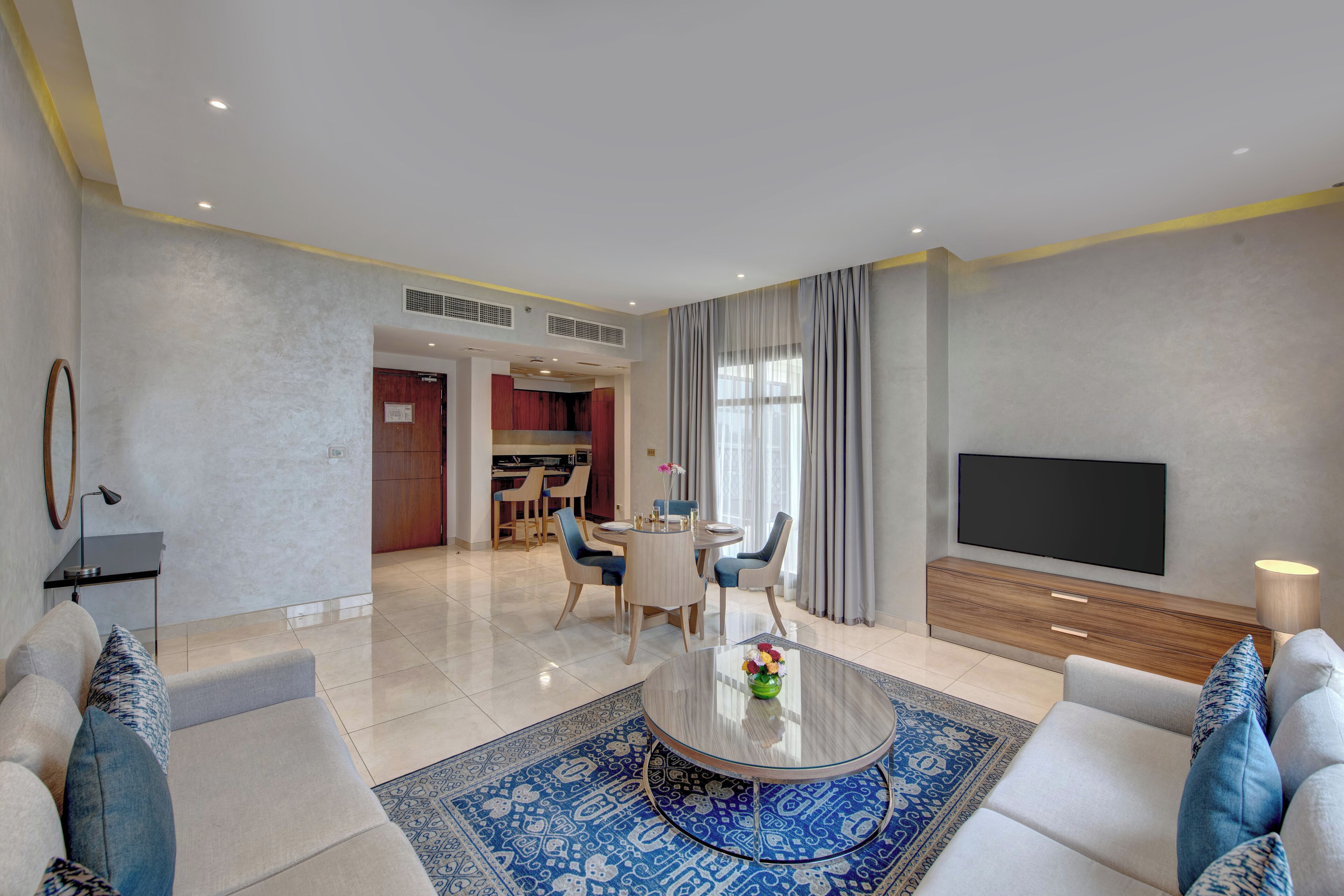 A modern living room with a sofa set, a coffee table with a vase, a flat-screen TV, and a dining area with chairs and a table.