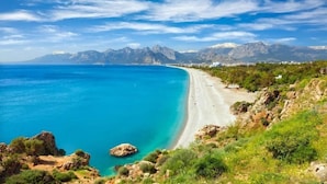 Un paisaje costero con una playa, afloramientos rocosos y montañas al fondo.