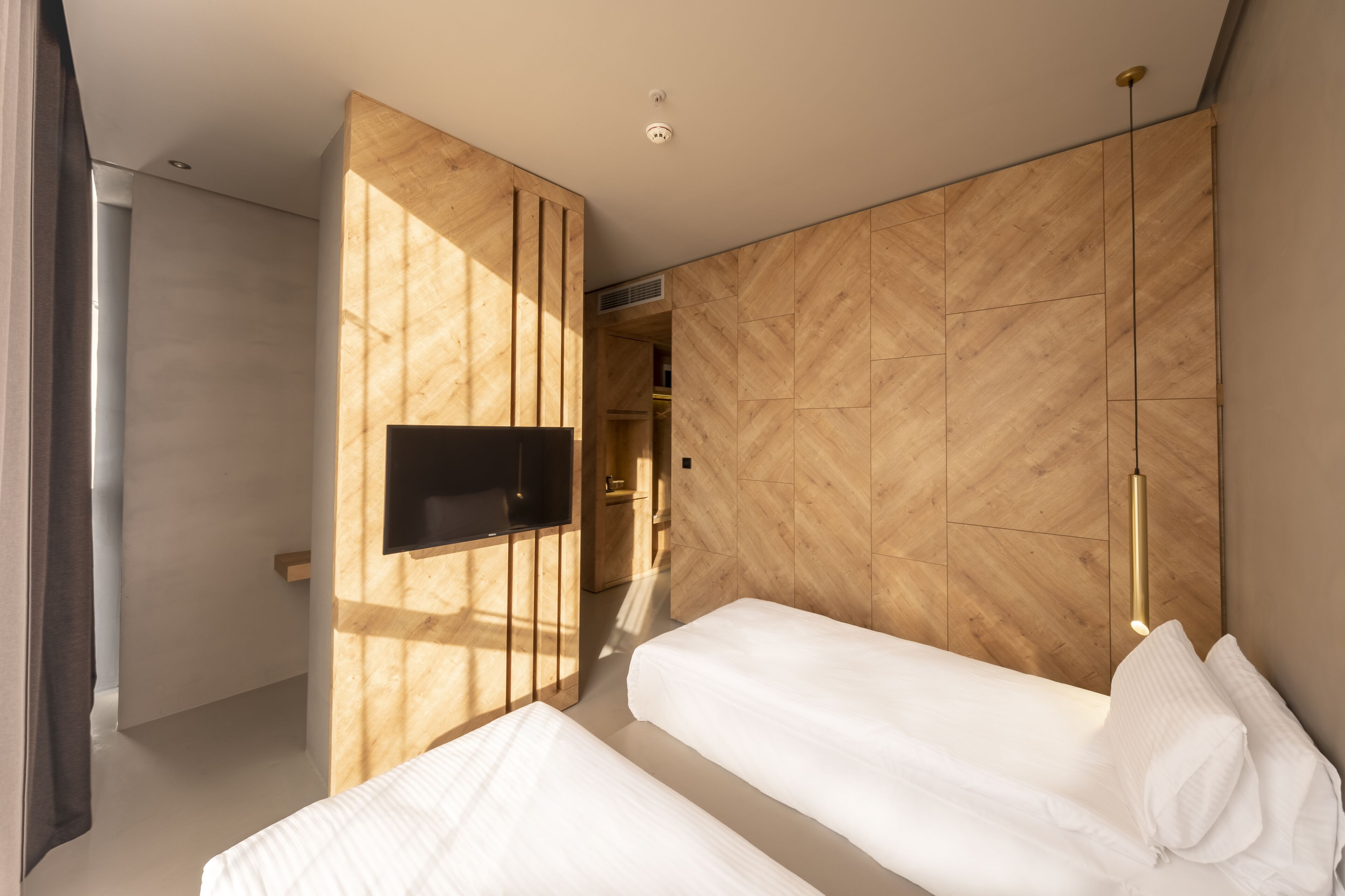 Una habitación de hotel moderna con dos camas, un televisor montado en un panel de madera y una lámpara de techo.