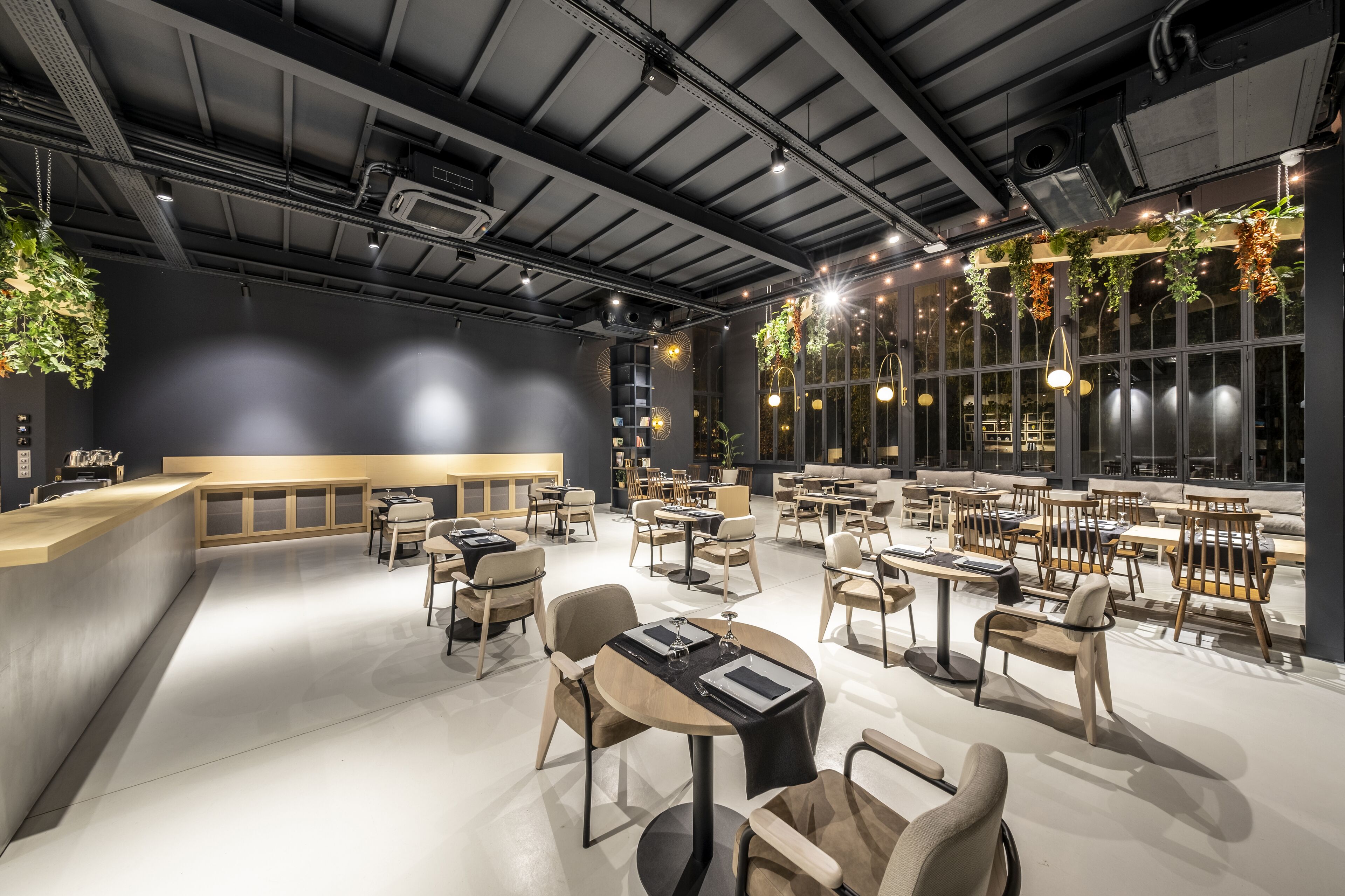 Un restaurant moderne avec un plafond haut, des poutres apparentes et des plantes suspendues. Les tables et les chaises sont disposées pour accueillir les clients.
