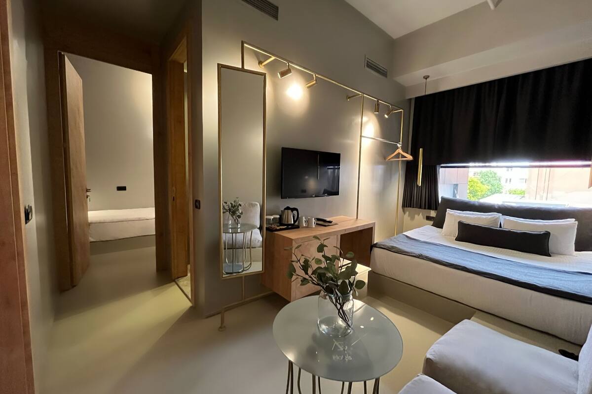 Ein modernes Hotelzimmer mit einem Bett, einem Sofa, einem kleinen Tisch mit einer Vase, einem Schreibtisch mit einem Fernseher und einem großen Fenster mit Vorhängen.