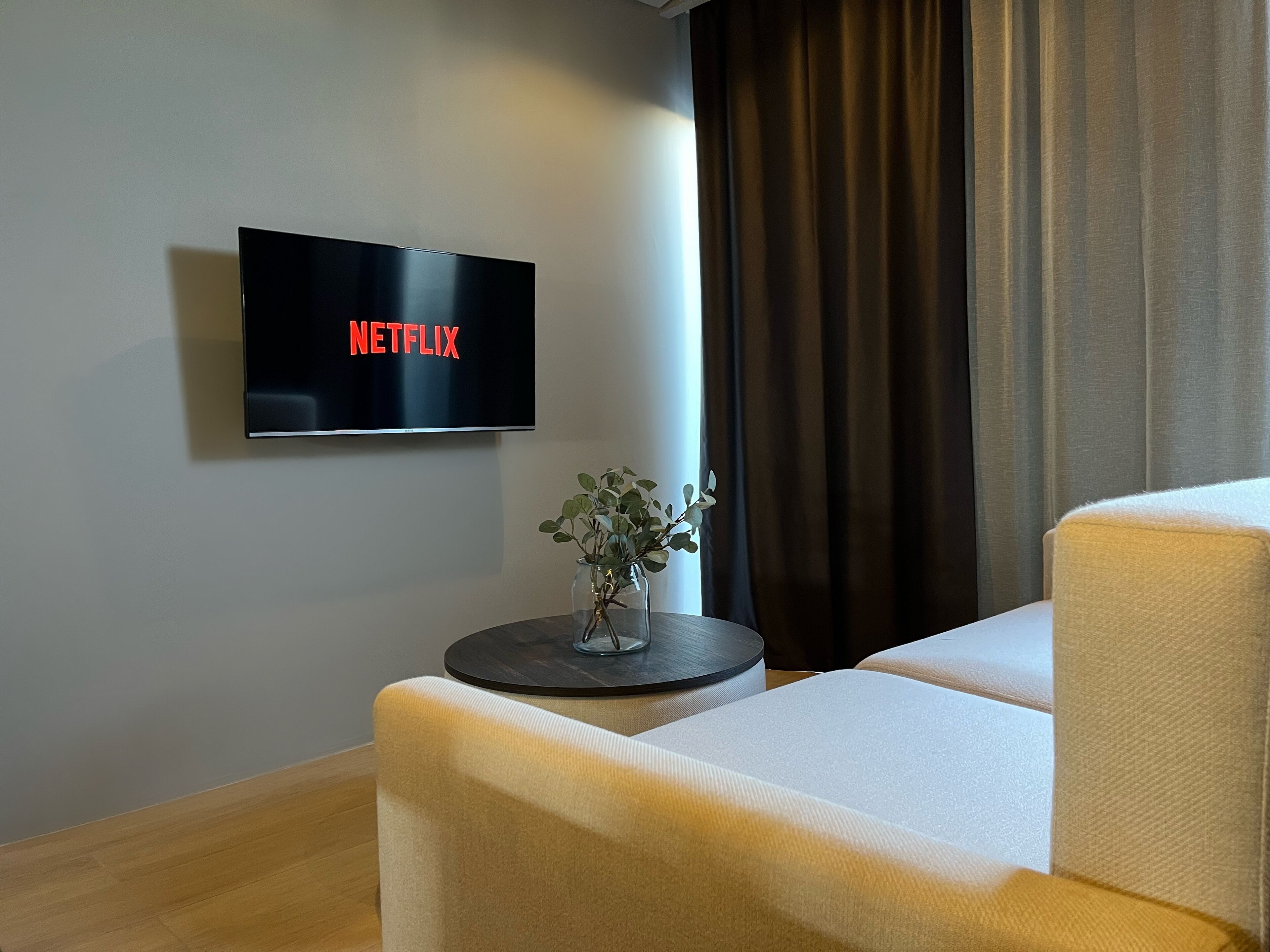 A modern living room with a flat-screen TV mounted on the wall displaying 'NETFLIX', a small round table with a vase of greenery, and a beige sofa.