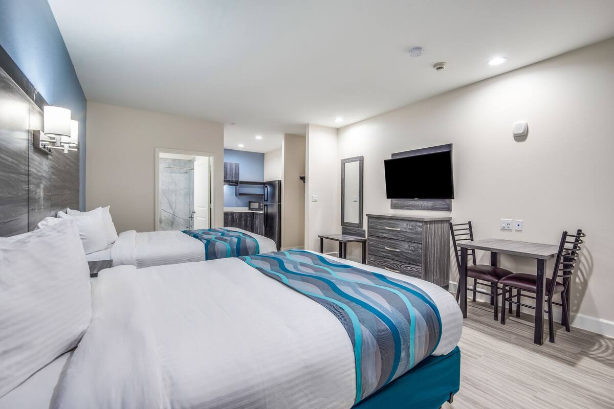 A hotel room with two beds, a desk, a chair, a TV, and a kitchenette in the background.