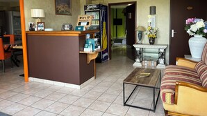 Un hall d’hôtel comprenant un comptoir d’accueil, une machine à café, un réfrigérateur, un canapé, une petite table et un vase avec des fleurs.