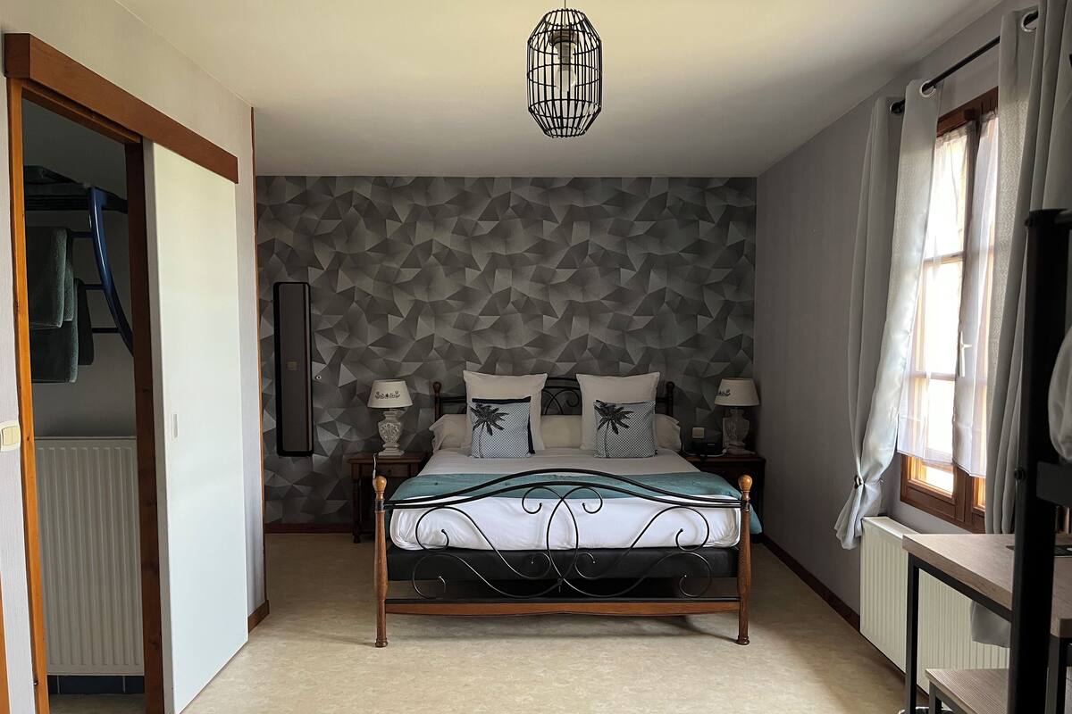 Une chambre avec un mur orné d’un motif géométrique, un lit avec un cadre en bois, deux tables de chevet avec des lampes, un luminaire suspendu et une fenêtre avec des rideaux.