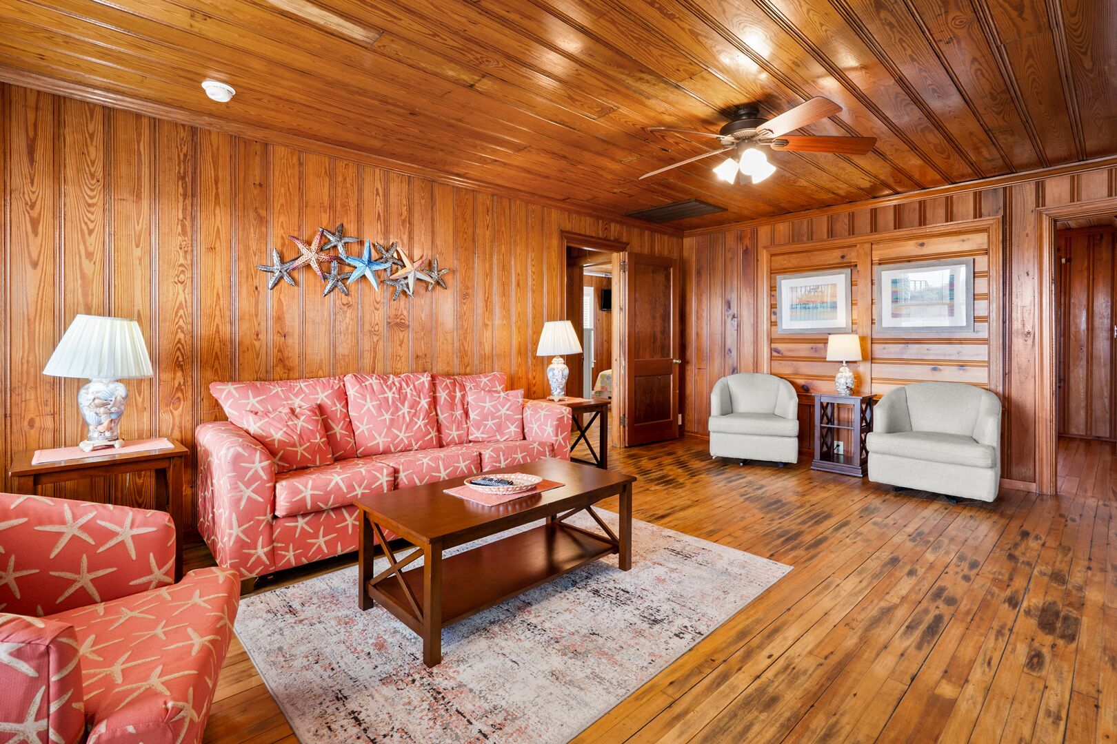 A wooden-paneled room with a red sofa, a wooden coffee table, two armchairs, and a ceiling fan.