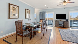 A dining area with a glass table and wooden chairs, a ceiling fan, a TV on a wooden cabinet, and a view of a body of water through a sliding glass door. - Stunning marsh view home, gorgeously furnished, private pool - walk to beach - perfect area (North Myrtle Beach)
