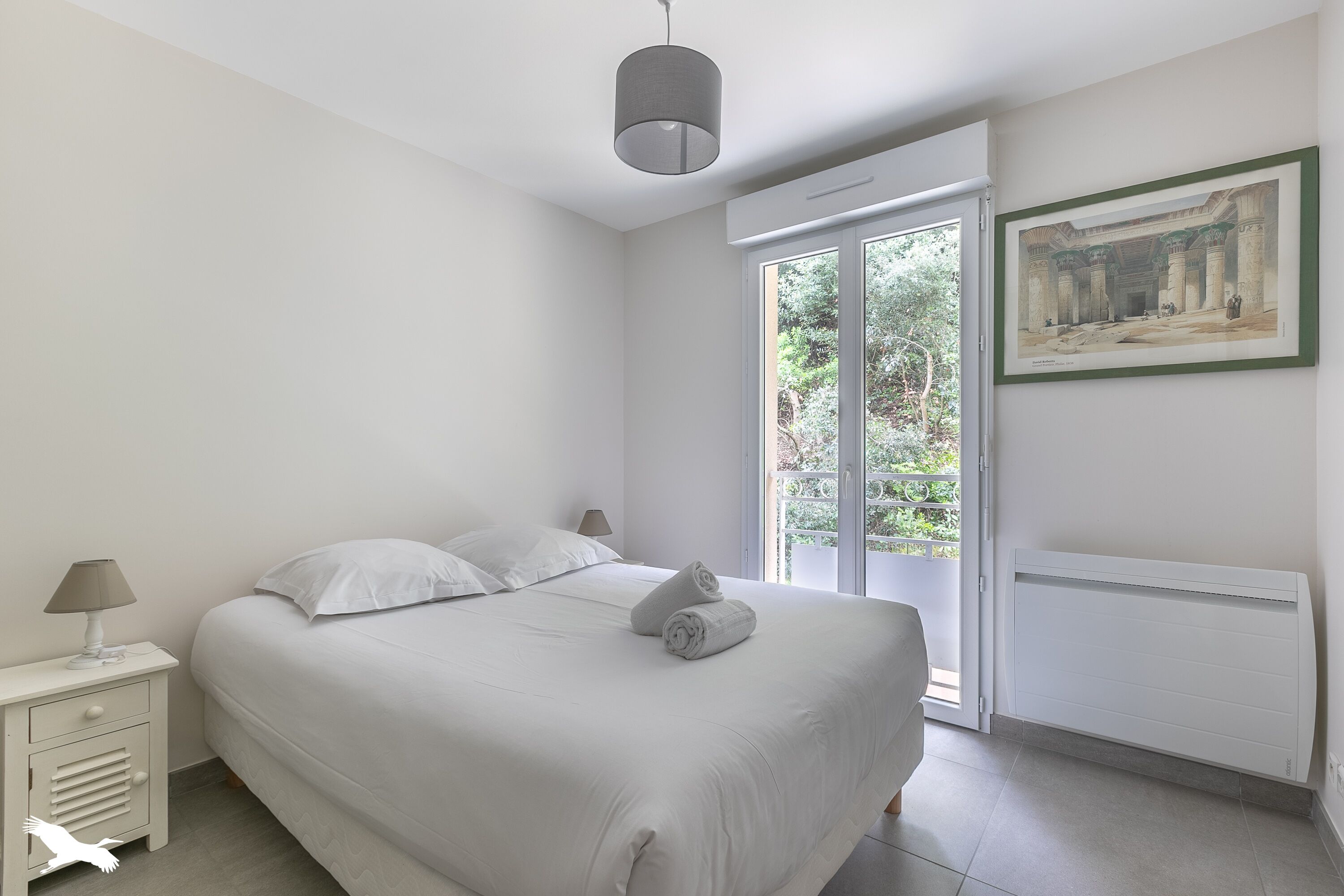 A bedroom with a bed, a nightstand, a lamp, a radiator, and a window with a view of greenery.