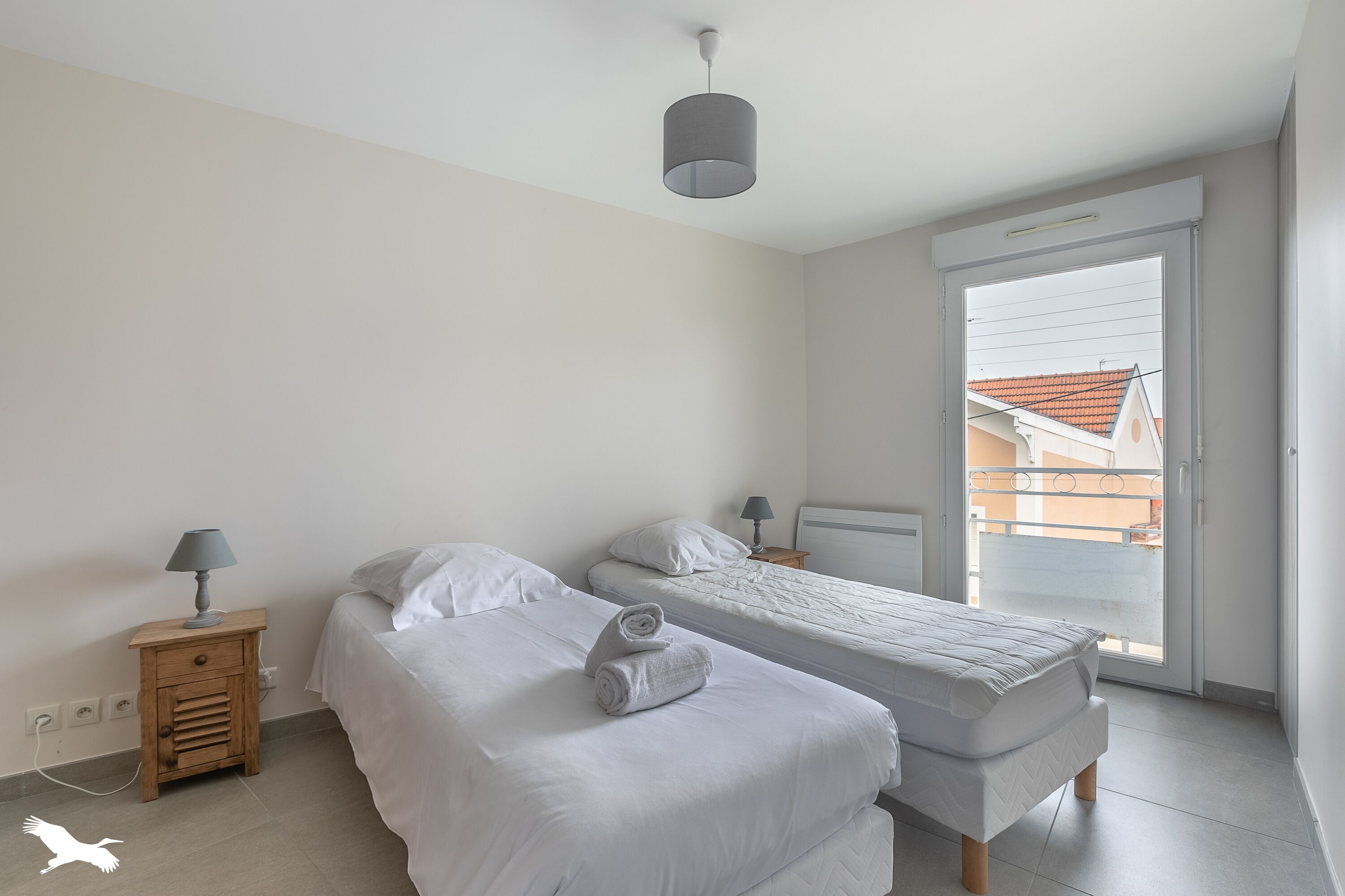 Quarto com duas camas, uma mesa de cabeceira de madeira, uma luminária e vista por uma porta de correr.