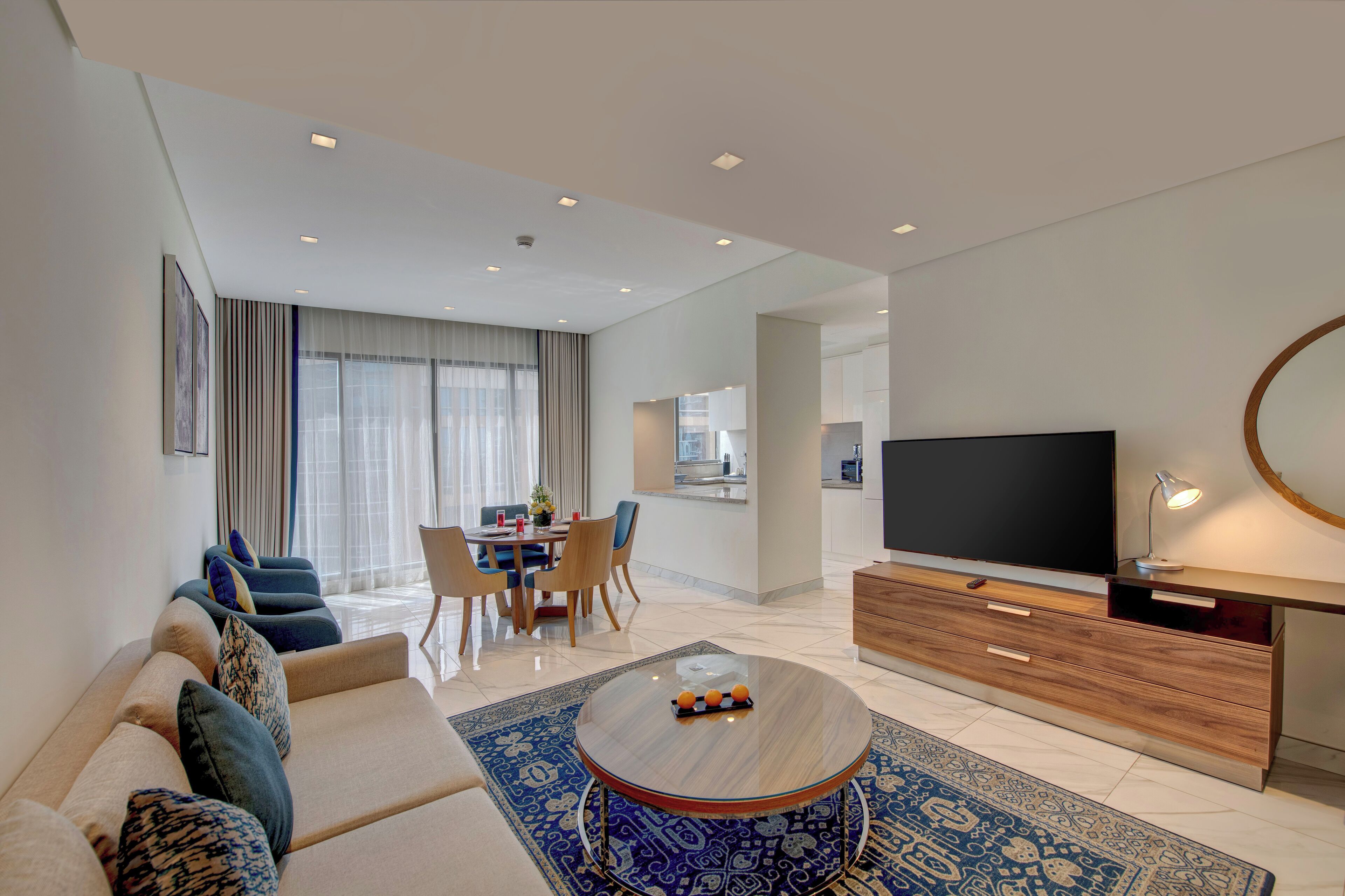 A modern living room with a sofa, coffee table, and TV. There is a dining area with chairs and a table. The room has a large window with curtains.