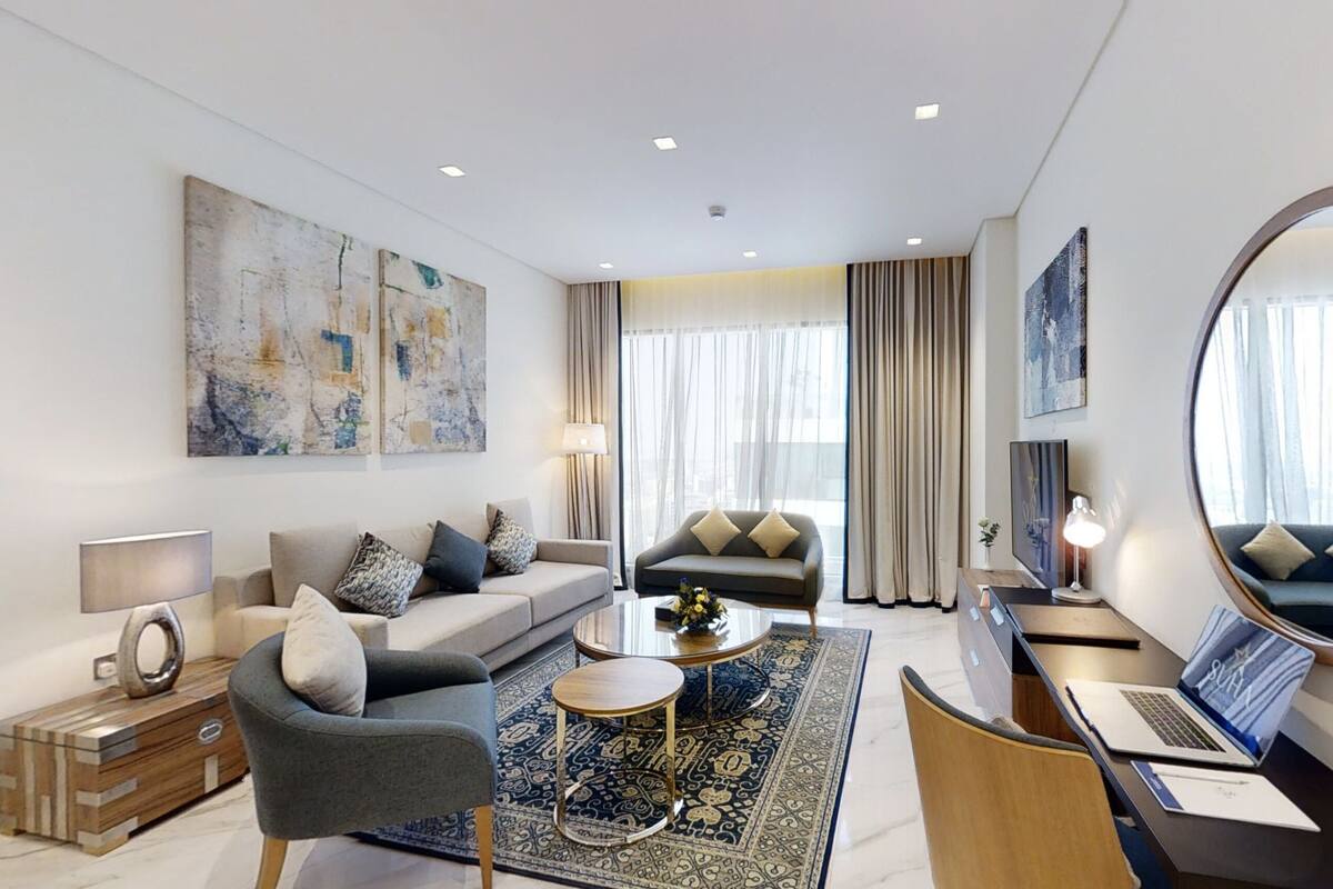 Une chambre d’hôtel moderne avec un canapé, un fauteuil et un bureau avec un ordinateur portable. On y trouve également un grand miroir, une lampe et un tapis décoratif.