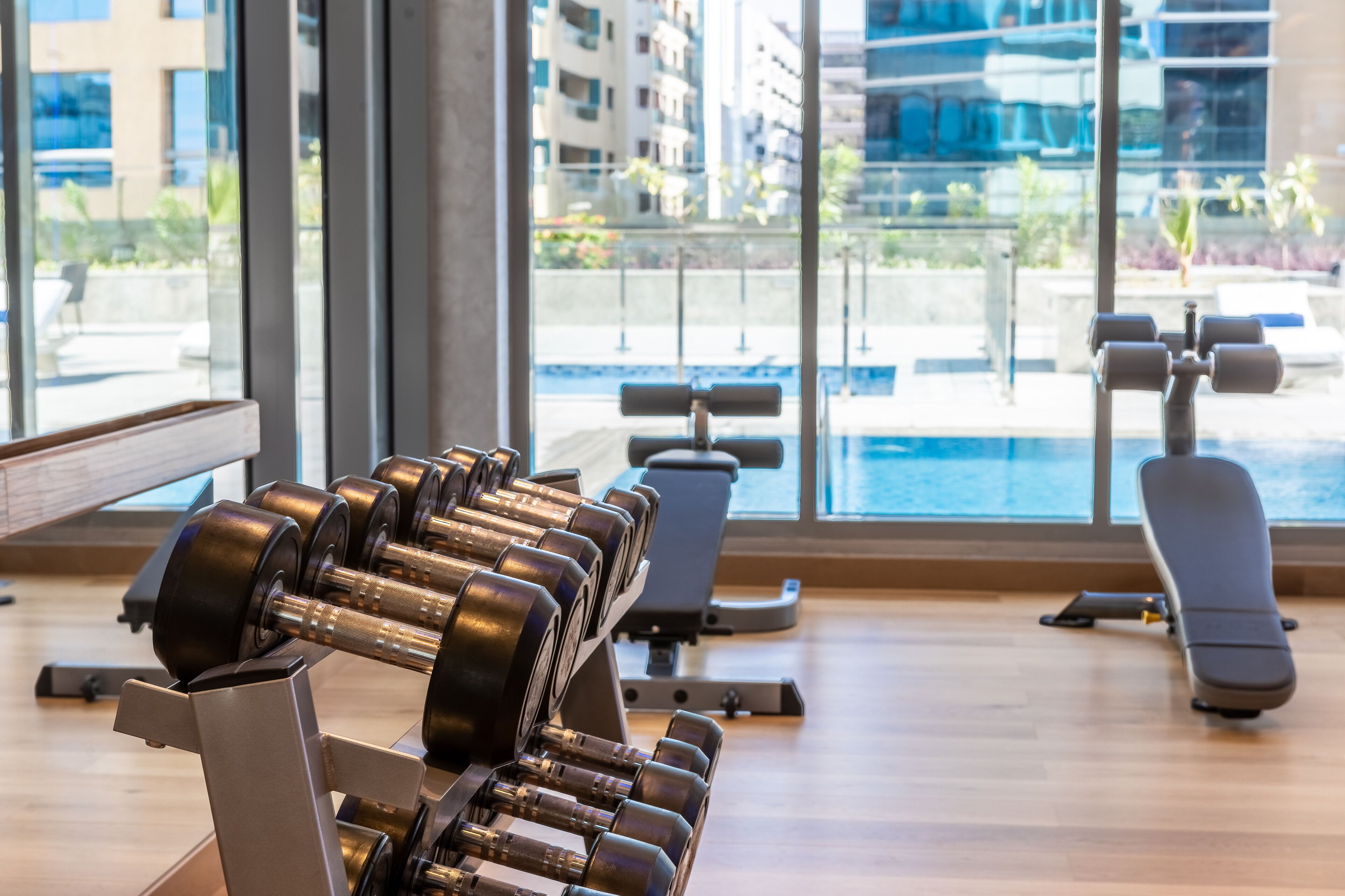 Ein Fitnessstudio mit Trainingsgeräten, großen Fenstern und Blick auf einen Pool sowie Gebäude.