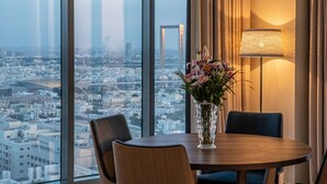 Hotelkamer met groot raam en uitzicht over de stad, een ronde tafel met twee stoelen en een vaas met bloemen.