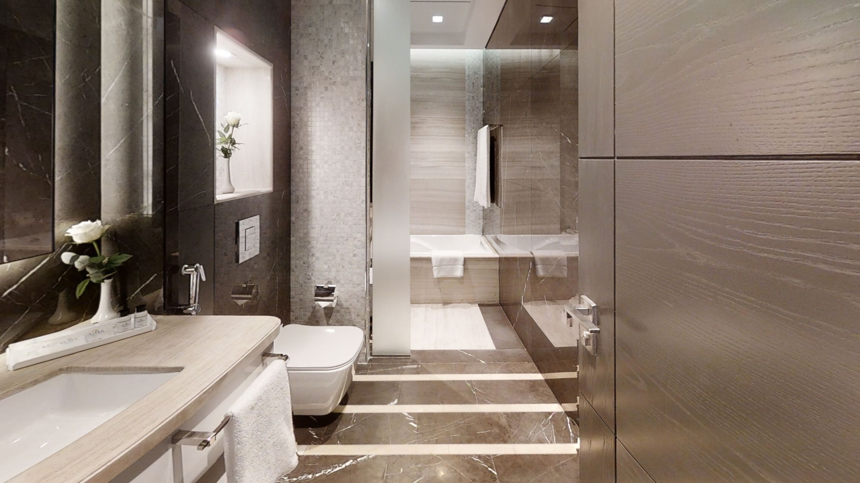 A modern bathroom with a marble floor, a large mirror, a white rose vase, and a glass shower enclosure.